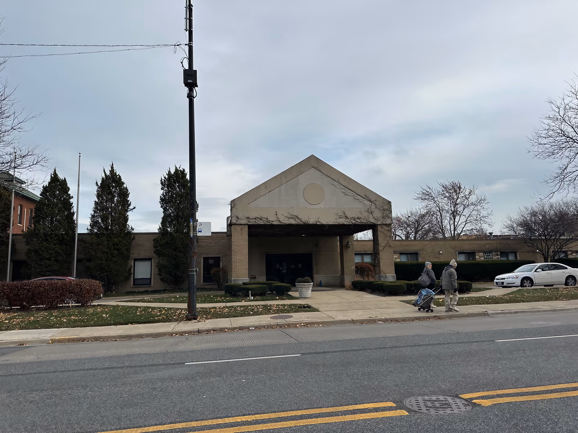 Exterior view of a single-story building with a peaked entrance canopy. Two people are walking on the sidewalk in front of the building, one pulling a cart. There are trees, bushes, and parked cars visible around the building under a cloudy sky.