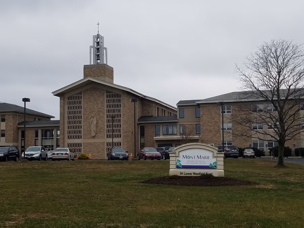 Exterior front view of Mont Marie Rehabilitation & Healthcare Center showing the brick building with a tower and cross, parking lot, and a facility sign on a grassy lawn.