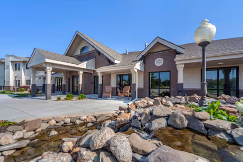 Exterior view of Valley Lakes Assisted Living facility showing a single-story building with a covered entrance, large windows, and a landscaped rock water feature in the foreground under a clear blue sky.
