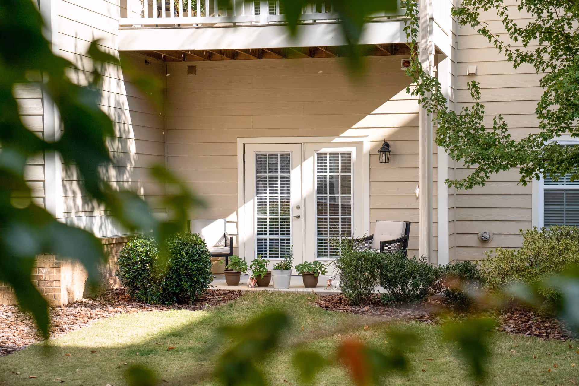 View of a patio area outside a senior living facility unit with two cushioned chairs, several potted plants, and bushes. The patio has double glass doors with white frames and blinds, and the building exterior is beige with white trim. Green leaves partially frame the image.