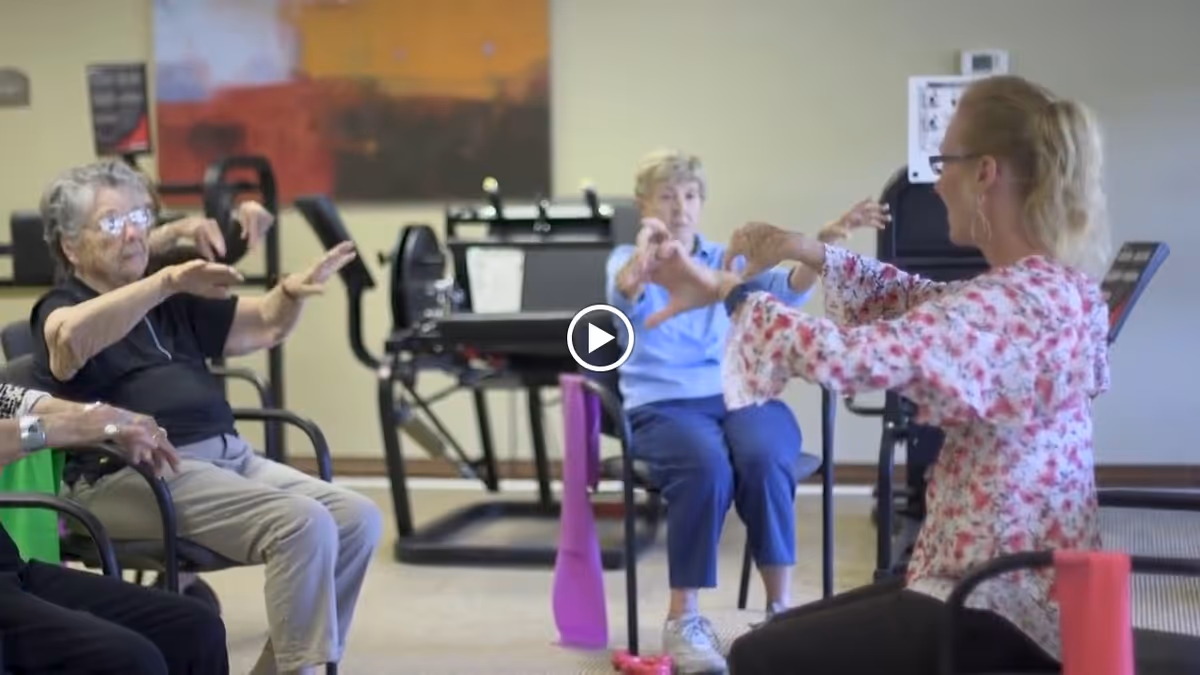 A group of elderly women seated in chairs participating in a seated exercise class led by an instructor in a floral blouse, with exercise equipment visible in the background.