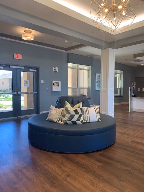Interior view of a senior living facility lobby with a round blue cushioned seating area in the center adorned with decorative pillows. The room has wooden flooring, gray walls, large windows, and a modern chandelier hanging from the ceiling. Double glass doors lead outside, and a reception desk is visible in the background with a person standing behind it.