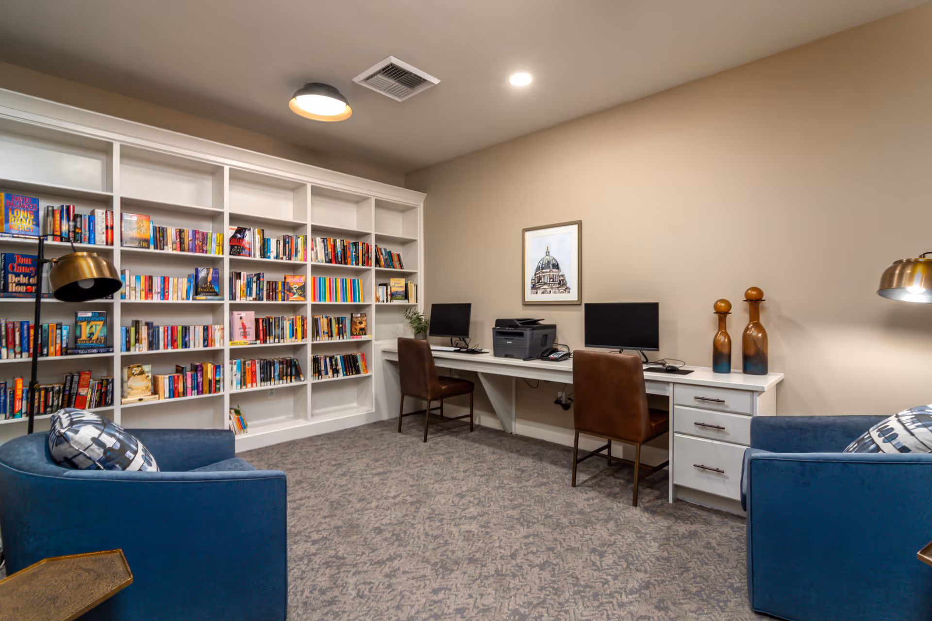 A cozy room with a large white bookshelf filled with books on the left wall, two blue armchairs with patterned pillows facing each other, and a long white desk against the right wall with two computer monitors, a printer, and two brown chairs. The room has beige walls, carpeted floor, and modern ceiling lighting.