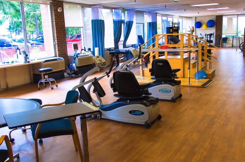 Bright rehabilitation gym with recumbent exercise bikes, parallel bars, therapy equipment and large windows.