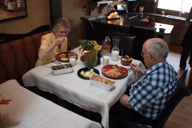 An elderly woman and man sit at a dining table in a kitchen area, eating a meal together. The table is covered with a white tablecloth and has various dishes, glasses of milk, and a potted plant as a centerpiece. The kitchen with countertops and appliances is visible in the background.