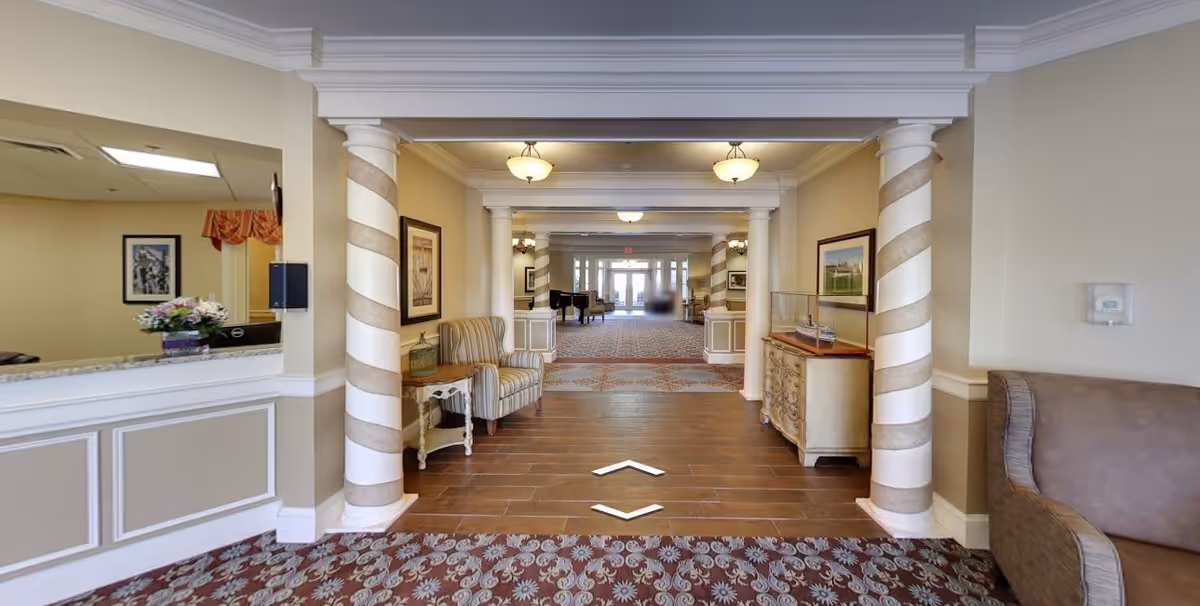 Interior view of a senior living facility lobby area with decorative striped columns, a striped armchair, a small table with a lamp, framed artwork on the walls, and a wooden sideboard. The floor transitions from patterned carpet to wood-like tiles leading to a larger common area with more seating and a piano in the distance.