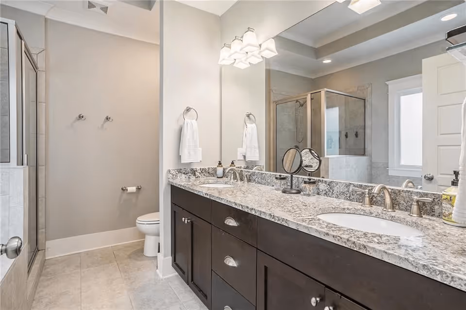 Modern bathroom with a double sink vanity featuring granite countertops and dark wood cabinets. There is a large mirror above the sinks with light fixtures mounted on the ceiling. To the left, there is a glass-enclosed shower and a toilet next to a towel rack with a white towel. The walls are painted light gray and the floor is tiled.