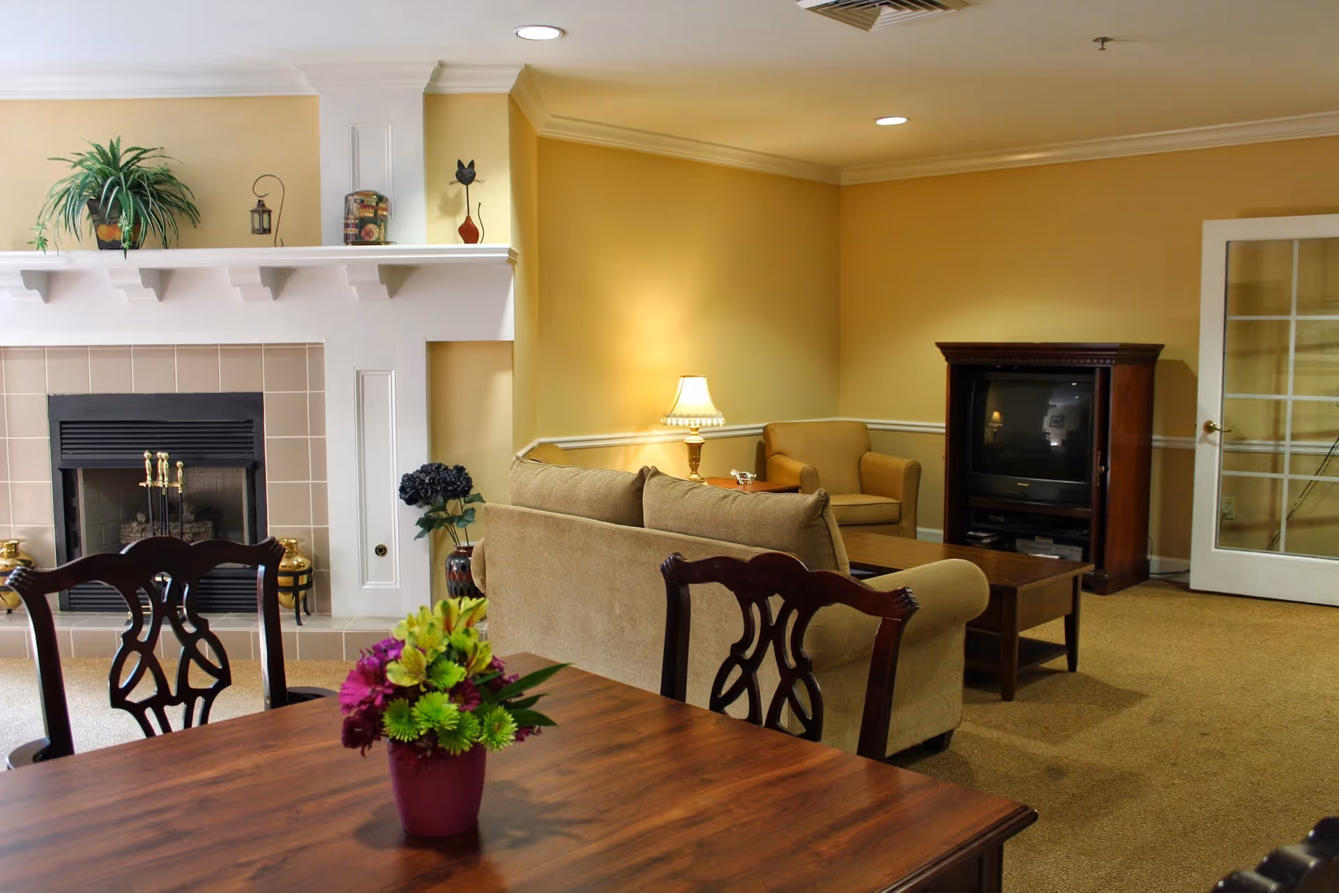Open living room and dining area with a fireplace, beige sofas, a wooden dining table with a flower arrangement, and a TV cabinet.