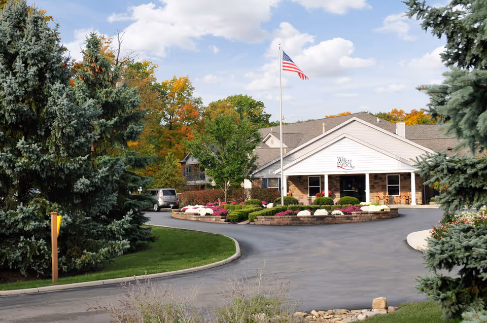 Exterior view of Willow Brook Christian Village facility with a circular driveway, landscaped flower beds, an American flag on a flagpole, and large evergreen trees on either side.