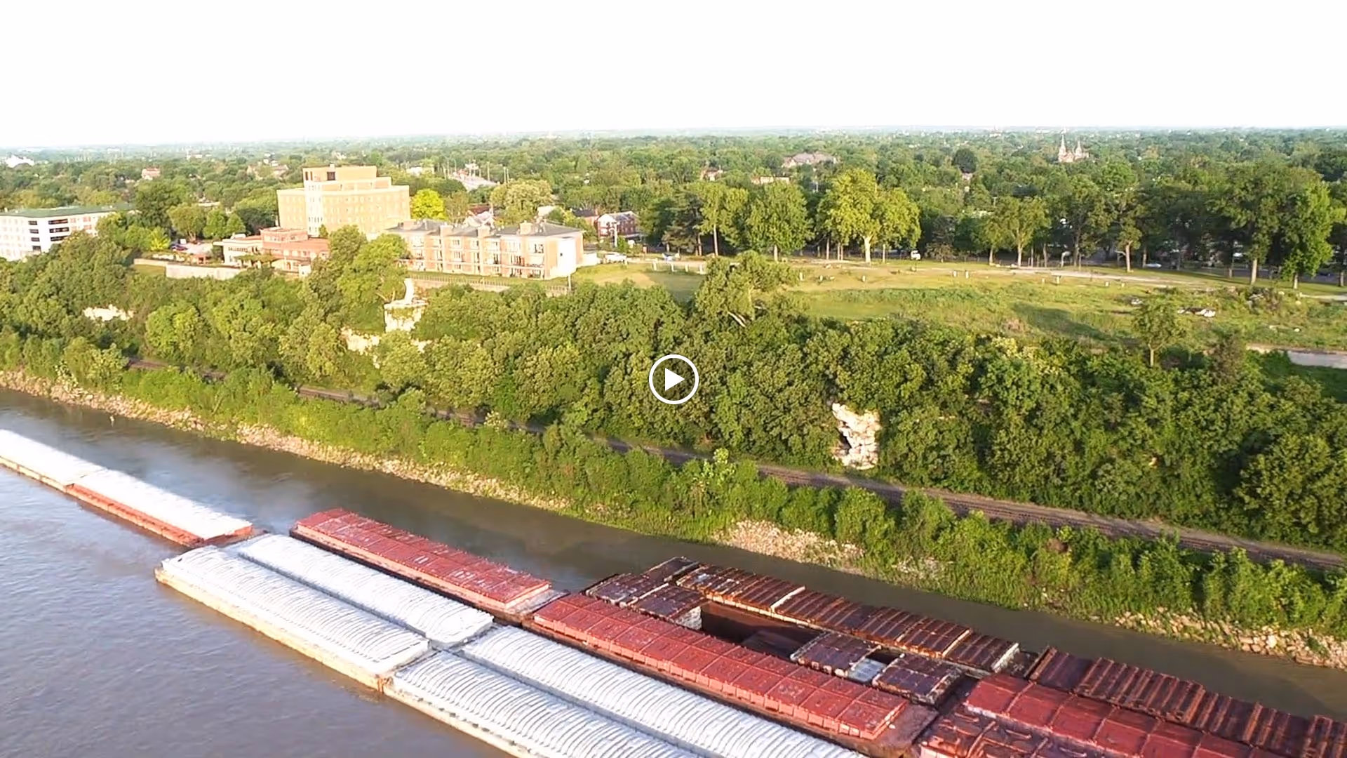 Aerial view of a river with barges docked along the shore, dense green trees lining the riverbank, and buildings visible in the background under a clear sky.