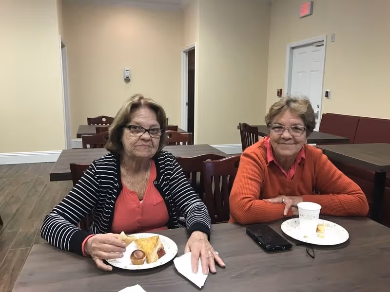 Two elderly women sitting at a wooden table in a dining area. One woman is wearing a black and white striped cardigan over a red top and is holding a piece of food. The other woman is wearing an orange sweater and glasses, with a cup and plate in front of her. The room has beige walls, wooden chairs, and tables, with a door and exit sign in the background.