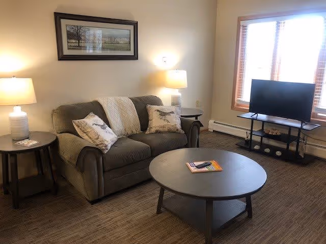 A cozy living room with a gray loveseat adorned with two decorative pillows and a throw blanket. There are two side tables with lamps on either side of the loveseat. A round coffee table with a remote control and a book is in front of the loveseat. A flat-screen TV is placed on a stand near a window with wooden blinds.