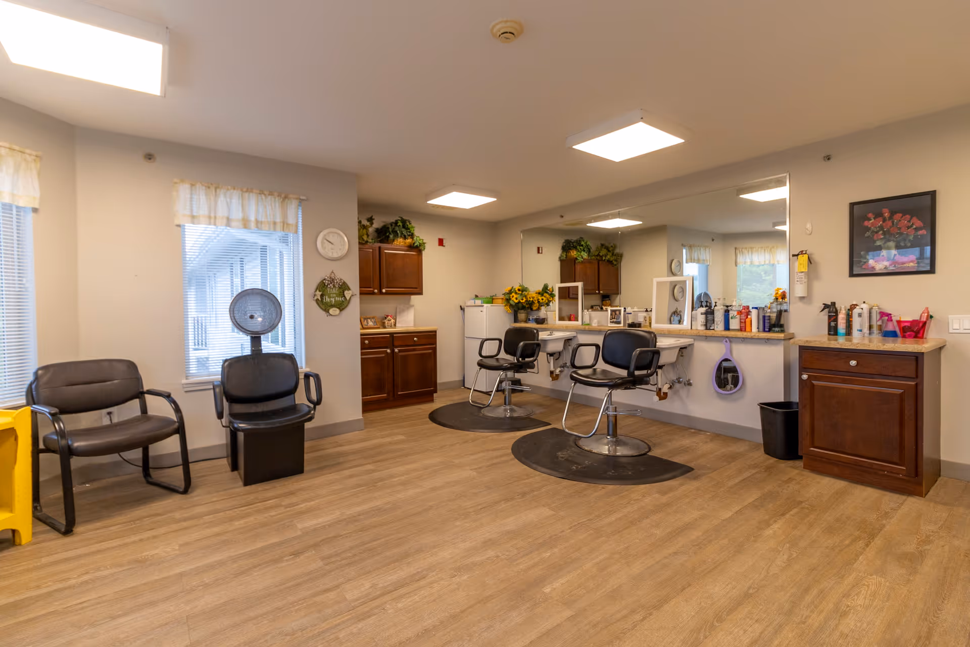 Interior view of a hair salon area in a senior living facility with two black salon chairs in front of a large mirror, a hair dryer chair, two waiting chairs, wooden cabinets, and various hair care products on the counter.