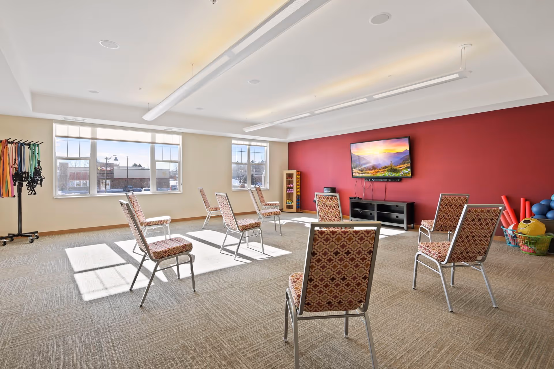 A bright, spacious room with large windows letting in natural light. The room has a red accent wall with a flat-screen TV mounted on it, and seven patterned chairs arranged in a circle on a carpeted floor. On the left side, there is a rack with colorful resistance bands, and on the right side, baskets with exercise balls and foam rollers are visible.