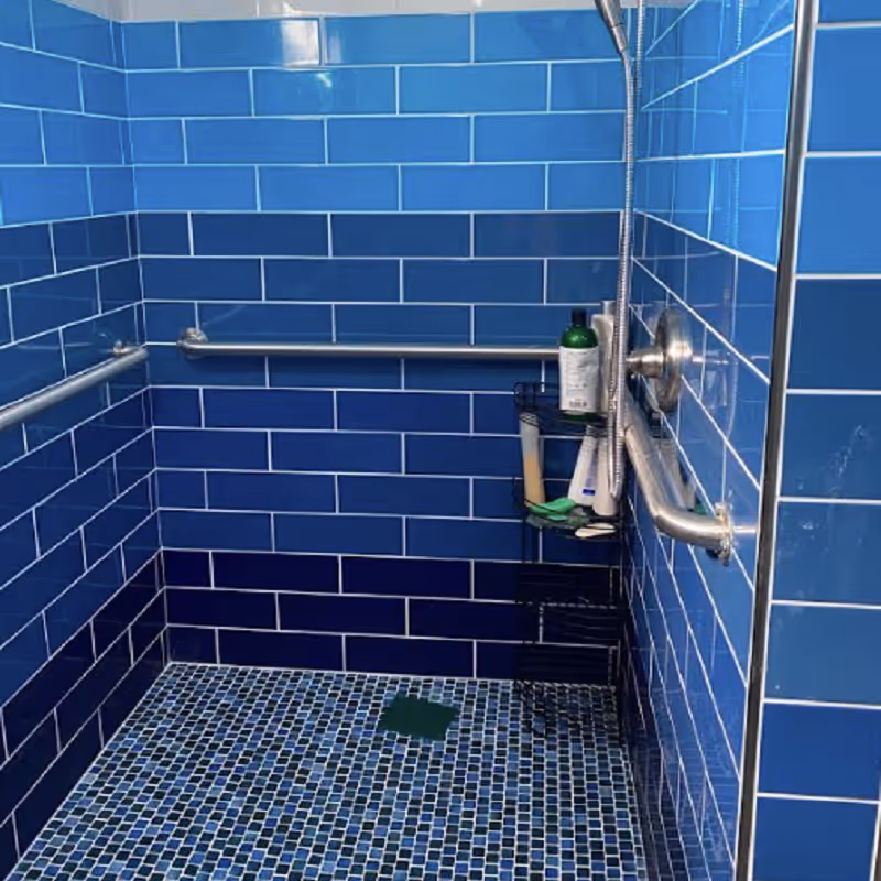 Blue tiled shower with grab bars, showerhead, and toiletries on a small wall rack.