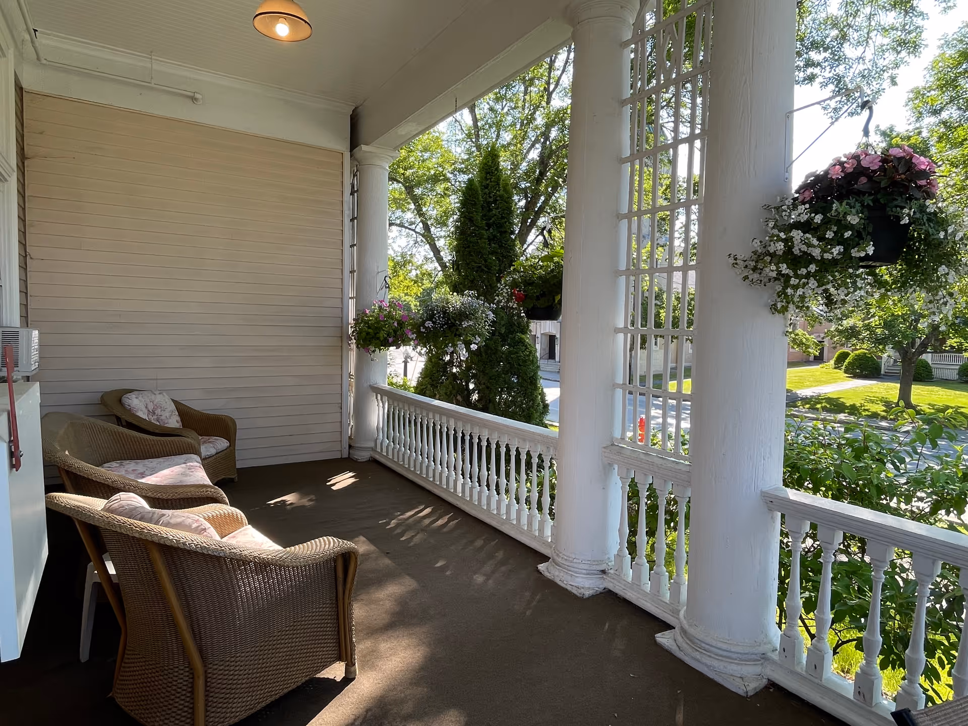 Shaded front porch with wicker chairs, white columns, hanging flower baskets, and a view of a tree-lined street.