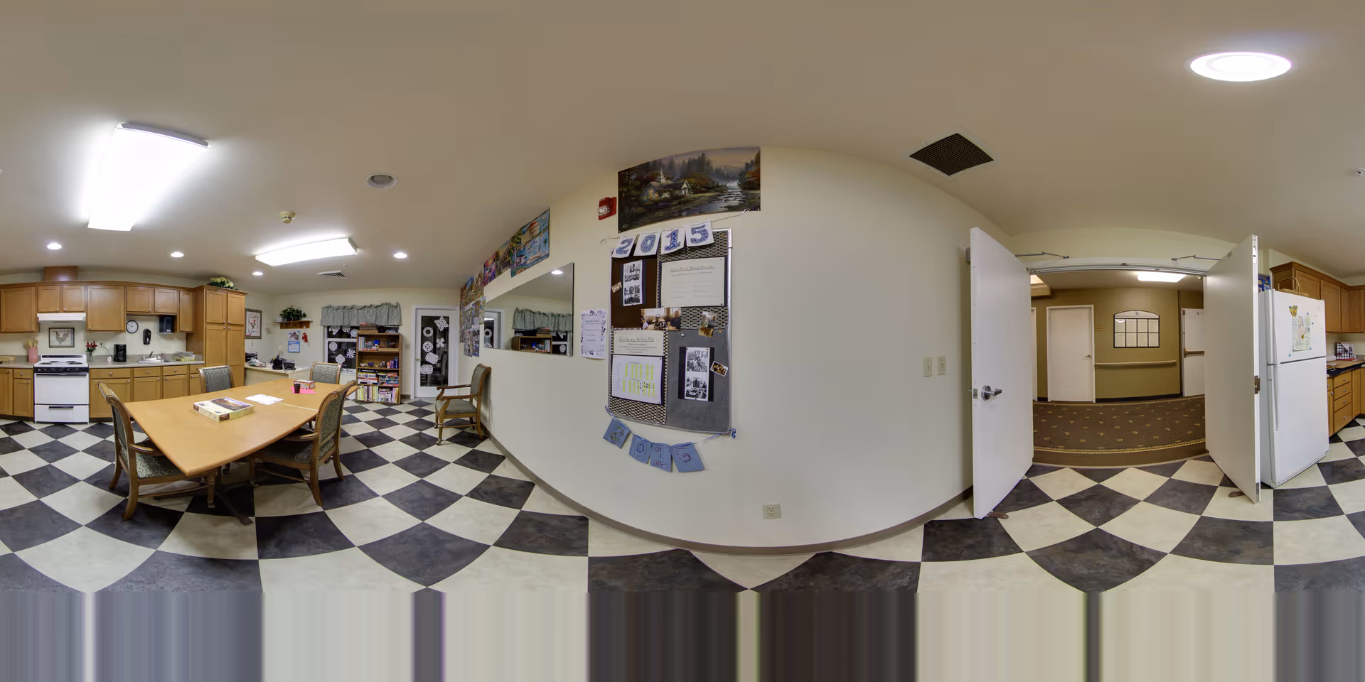 A communal kitchen and dining area in a senior living facility with a checkered black and white tile floor. The room features wooden cabinets, a white stove, a refrigerator, a wooden dining table with chairs, and a bulletin board on the wall. There is an open doorway leading to a carpeted hallway.