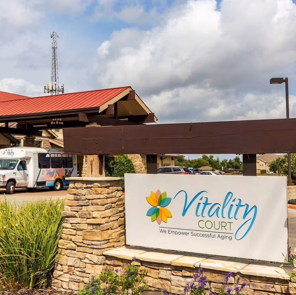Entrance sign for Vitality Court, a senior living facility, displayed on a stone base with a colorful flower logo and the tagline 'We Empower Successful Aging'. In the background, there is a building with a red roof, a parking lot with cars, and a shuttle bus with the Vitality Court logo.