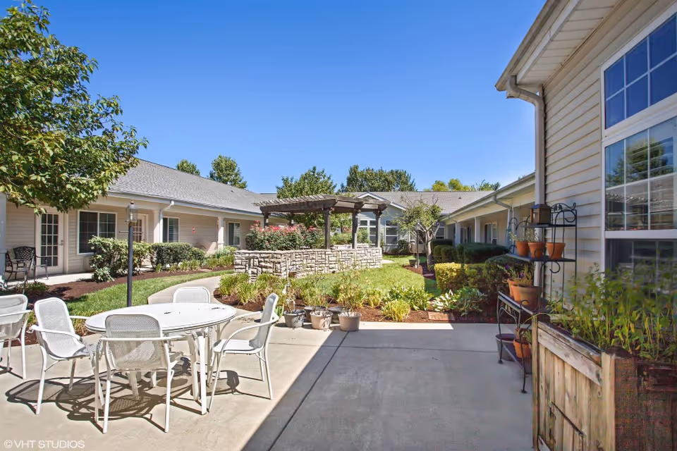 Outdoor courtyard area of a senior living facility with a round white table and chairs on a concrete patio, surrounded by well-maintained landscaping including bushes, trees, and potted plants. There is a stone planter with a wooden pergola in the center, and single-story building wings with windows and doors enclosing the courtyard under a clear blue sky.