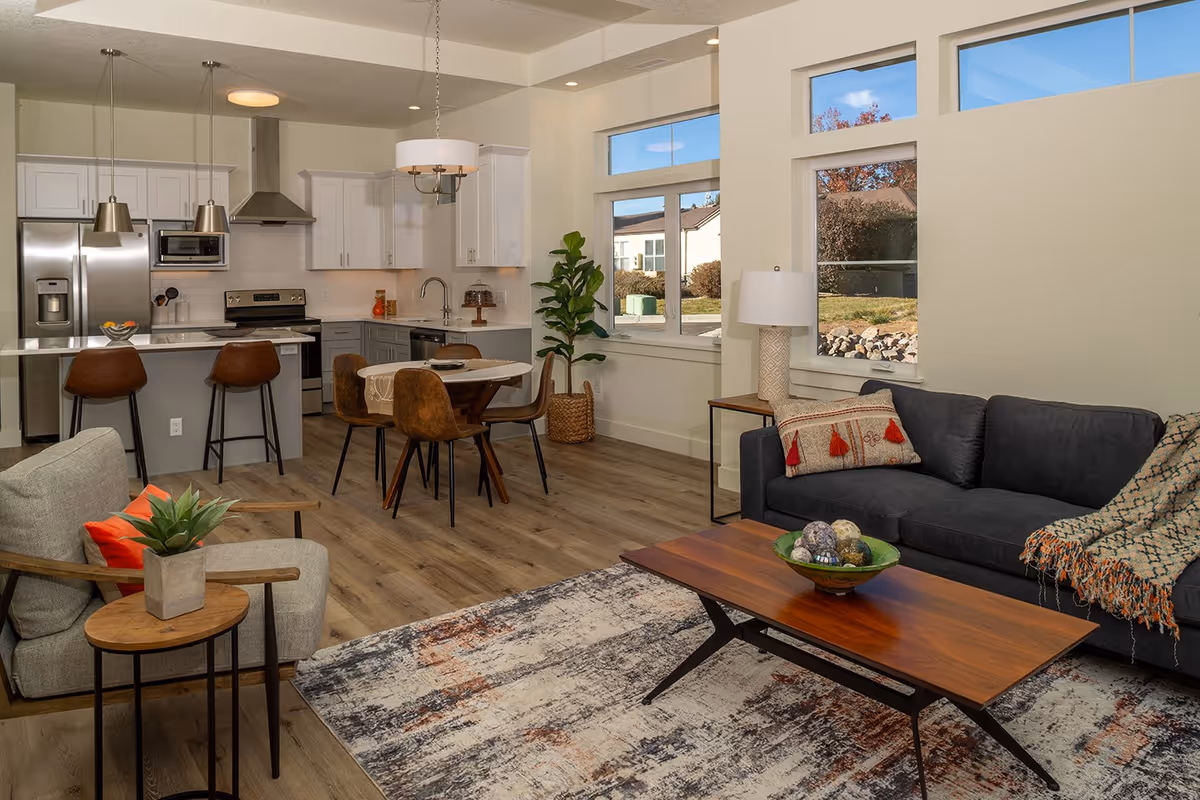 Open concept living space featuring a modern kitchen with white cabinets, stainless steel appliances, and a breakfast bar with two brown stools. Adjacent dining area with a round table and four brown chairs. Living room area with a dark gray sofa adorned with pillows and a throw blanket, a wooden coffee table with decorative items, a gray armchair with a red pillow, and a small side table with a plant. Large windows allow natural light to fill the room, showing an outdoor view of neighboring houses and greenery.
