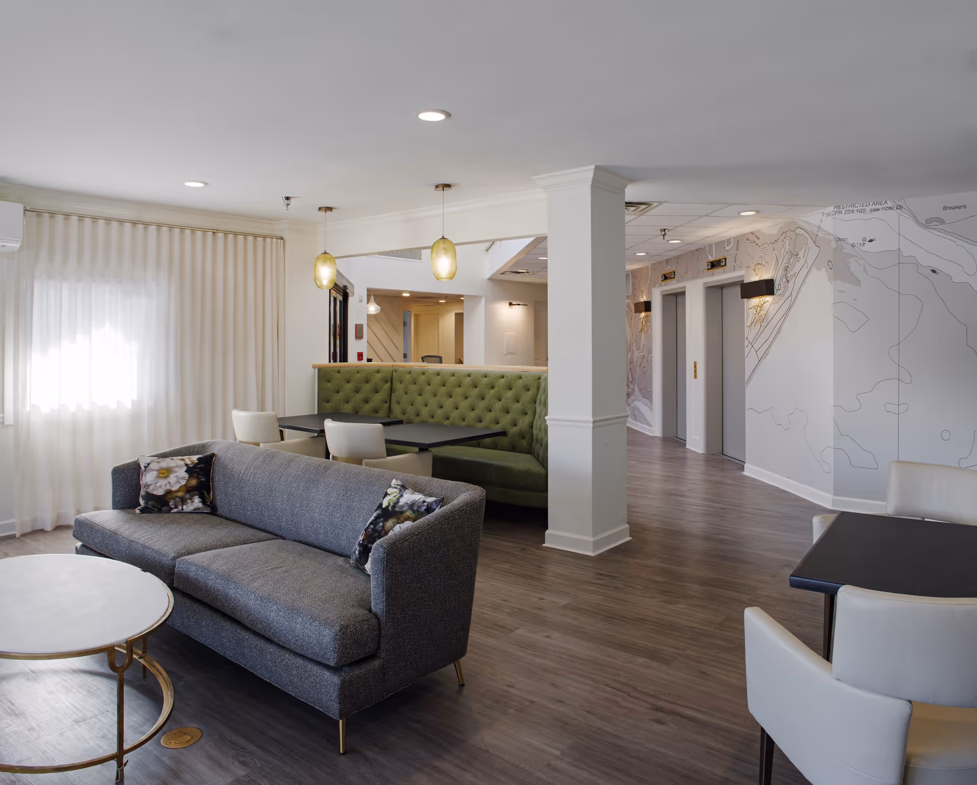 A modern and bright common area in a senior living facility featuring a gray sofa with floral pillows, a round white coffee table with gold legs, a green tufted booth seating area with black tables, white chairs, wood flooring, and two elevators in the background with wall sconces and a map design on the wall.