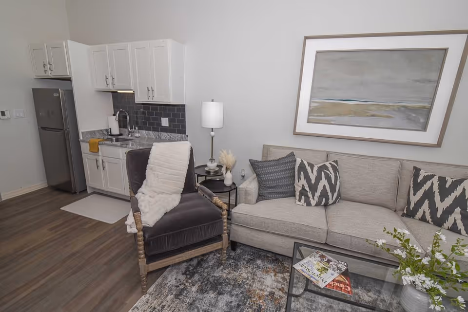 Open living area with a sofa, patterned pillows, an armchair and coffee table beside a small kitchenette with refrigerator and cabinets.