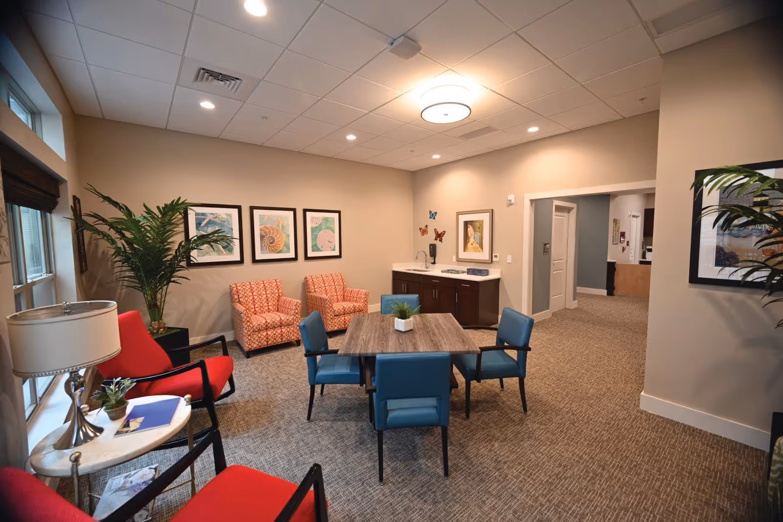 A cozy senior living common area with a square wooden table surrounded by four blue chairs. There are two red patterned armchairs and two solid red chairs near a window with a small round table holding a lamp and some items. The walls are beige with framed artwork and butterfly decorations. A small sink and cabinet area is visible in the corner, and a hallway leads to other rooms.