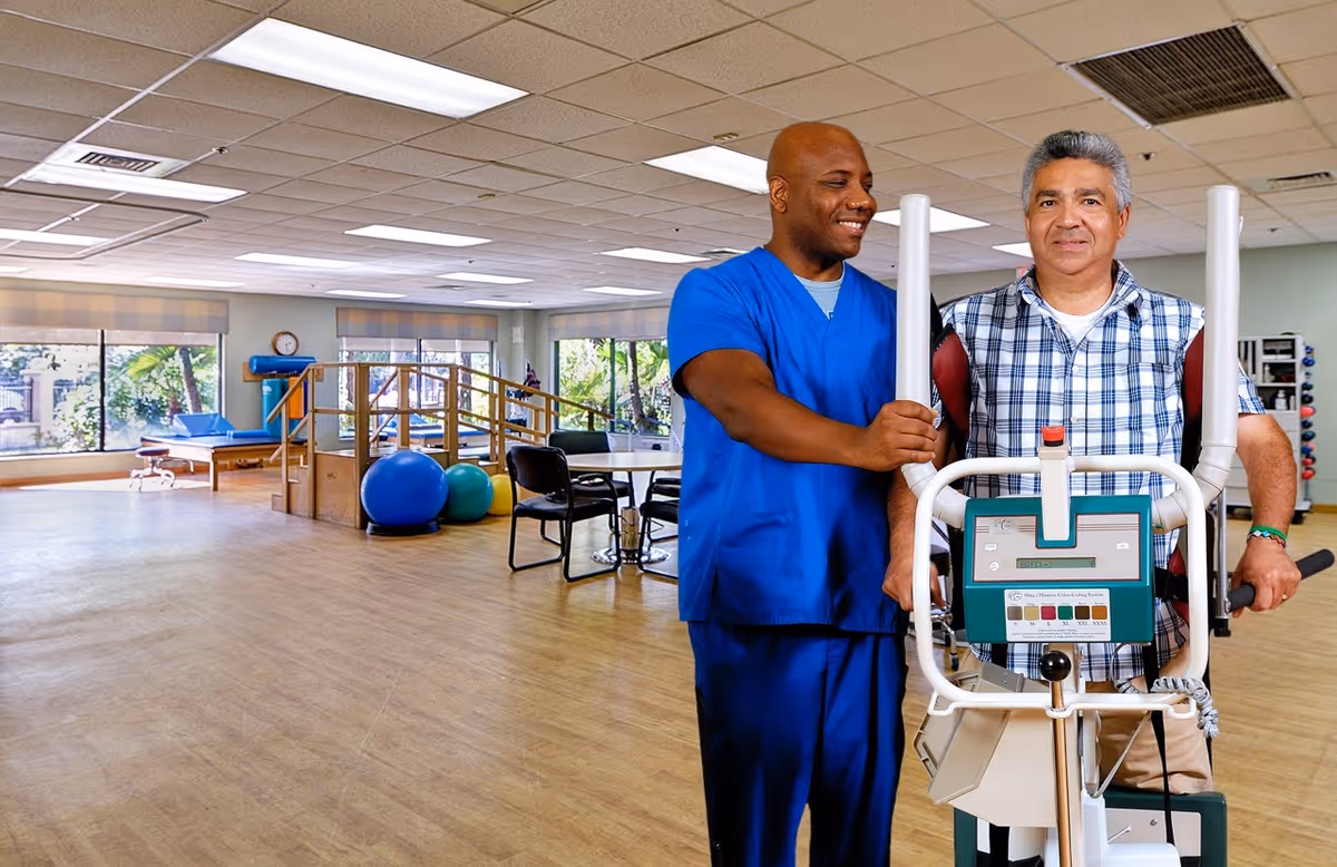 A healthcare professional in blue scrubs assists an older man using a piece of exercise equipment in a spacious physical therapy or rehabilitation room with large windows, exercise balls, and therapy tables.