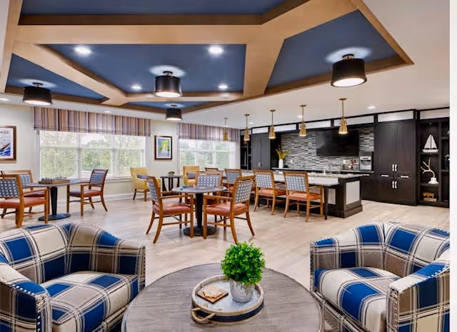 A spacious and well-lit common area with a modern kitchen and dining space. The room features a ceiling with wooden beams and multiple black light fixtures. There are several tables with chairs arranged for dining, and a kitchen island with bar stools. In the foreground, two blue and white plaid armchairs surround a round wooden coffee table with a small green plant and decorative items. Large windows with striped valances allow natural light to fill the room.
