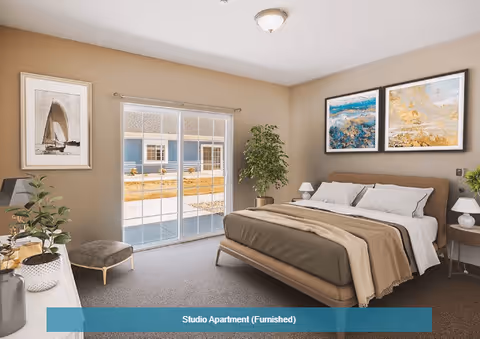 A furnished studio apartment bedroom with a large bed covered in beige and brown bedding, two framed abstract paintings above the bed, a nightstand with a lamp on each side, a potted plant in the corner, a small ottoman near a sliding glass door that leads outside, and a dresser with decorative items on the left side.