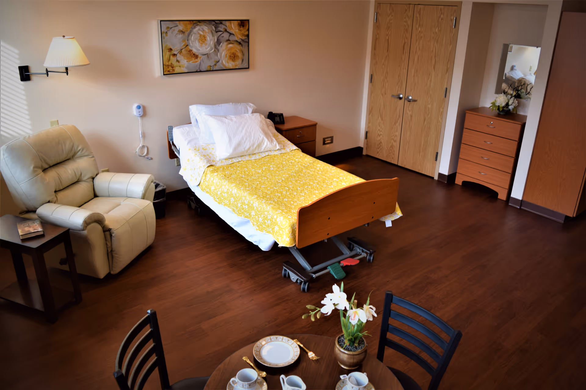 A cozy personal care room with a single adjustable bed covered with a yellow floral blanket, a beige recliner chair next to a small side table with a book, a wooden dresser with a mirror and flowers, a double-door closet, and a round dining table set with plates, cups, and a flower pot. The room has wooden flooring and soft lighting.