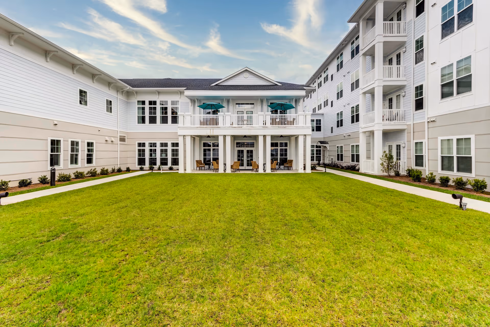 A green lawn courtyard framed by a multi-story white senior living building with balconies and a covered patio.