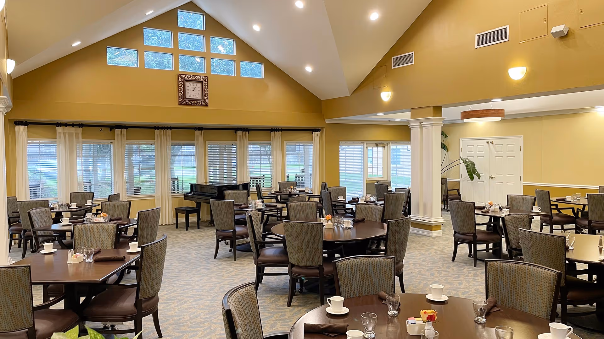 A spacious dining room with multiple round tables set with cups, glasses, and napkins. The room features high vaulted ceilings with recessed lighting, large windows with white curtains, and a grand piano near the back wall. The walls are painted a warm yellow color, and there is a large clock mounted above the windows.