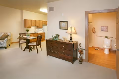 Interior view of a senior living facility apartment showing a small dining area with a round table and four chairs, a kitchenette with wooden cabinets, a microwave, and a countertop. To the right, there is a dresser with a table lamp and decorative items. An open door reveals a bathroom with a toilet, grab bars, and a framed picture on the wall.