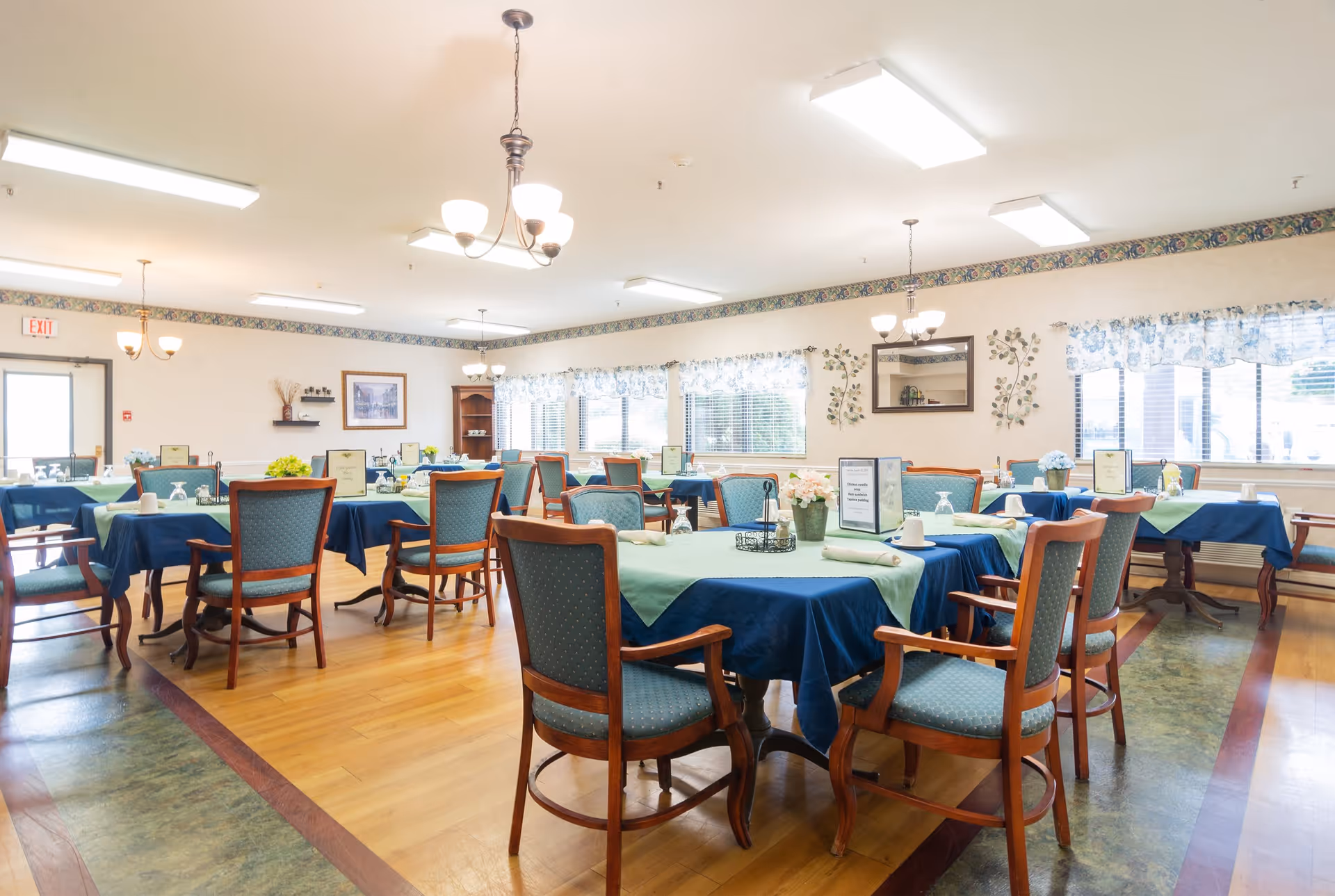 A bright and spacious dining room with multiple tables covered in blue and green tablecloths, each set with cups, glasses, and napkins. Wooden chairs with green upholstery surround the tables. The room has large windows with floral valances, light wood flooring, and decorative wall art. Ceiling lights and chandeliers provide ample lighting.
