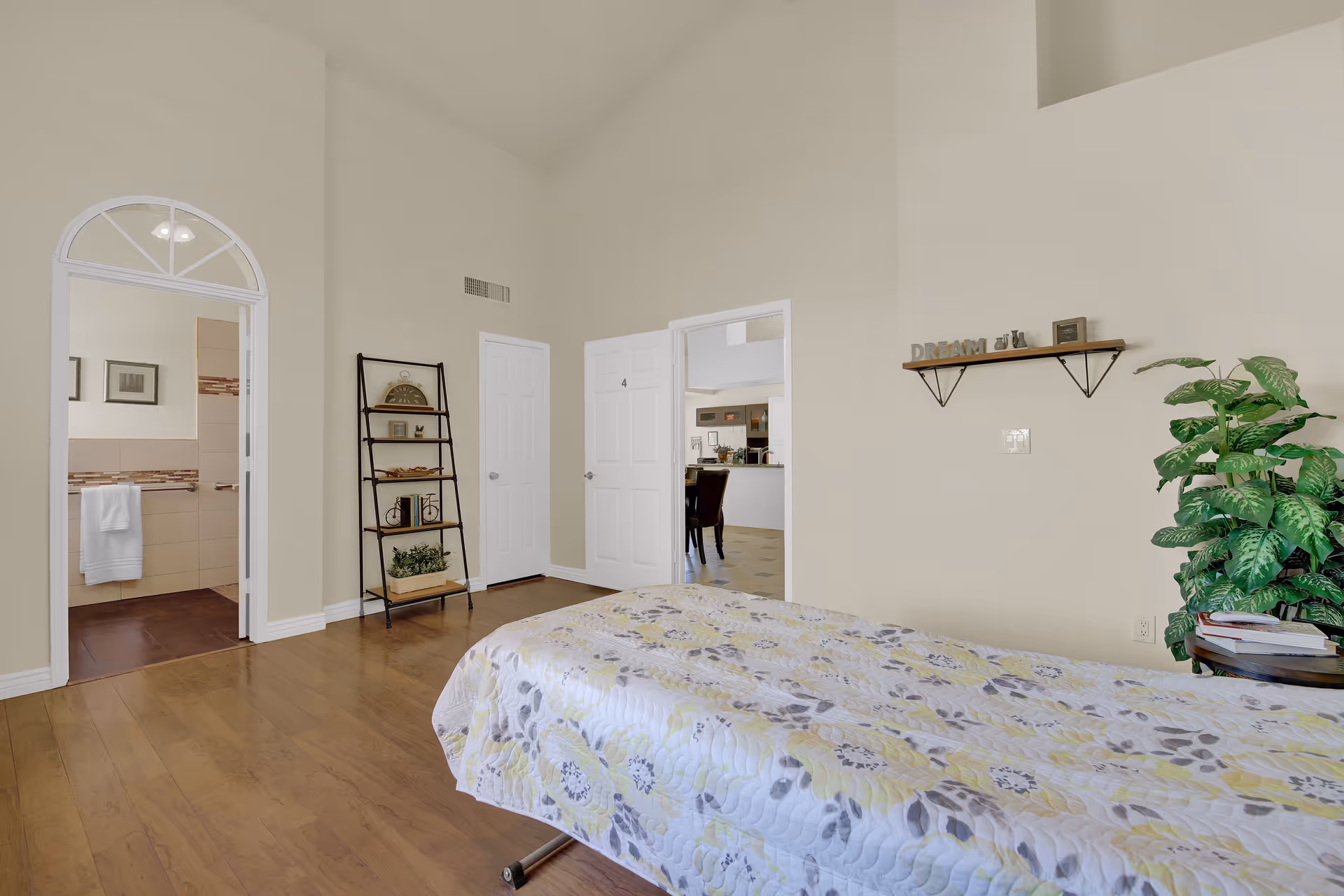 A bedroom with a bed covered in a floral quilt, wooden flooring, a small round table with books and a plant, a wall shelf with decorative items and the word 'DREAM', a black metal ladder-style shelf with books and decor, and two white doors leading to other rooms including a bathroom.