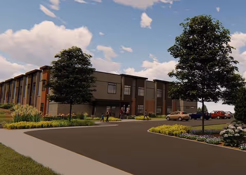 Two-story modern senior living building with a landscaped driveway, trees, parked cars, and people near the entrance under a partly cloudy sky.