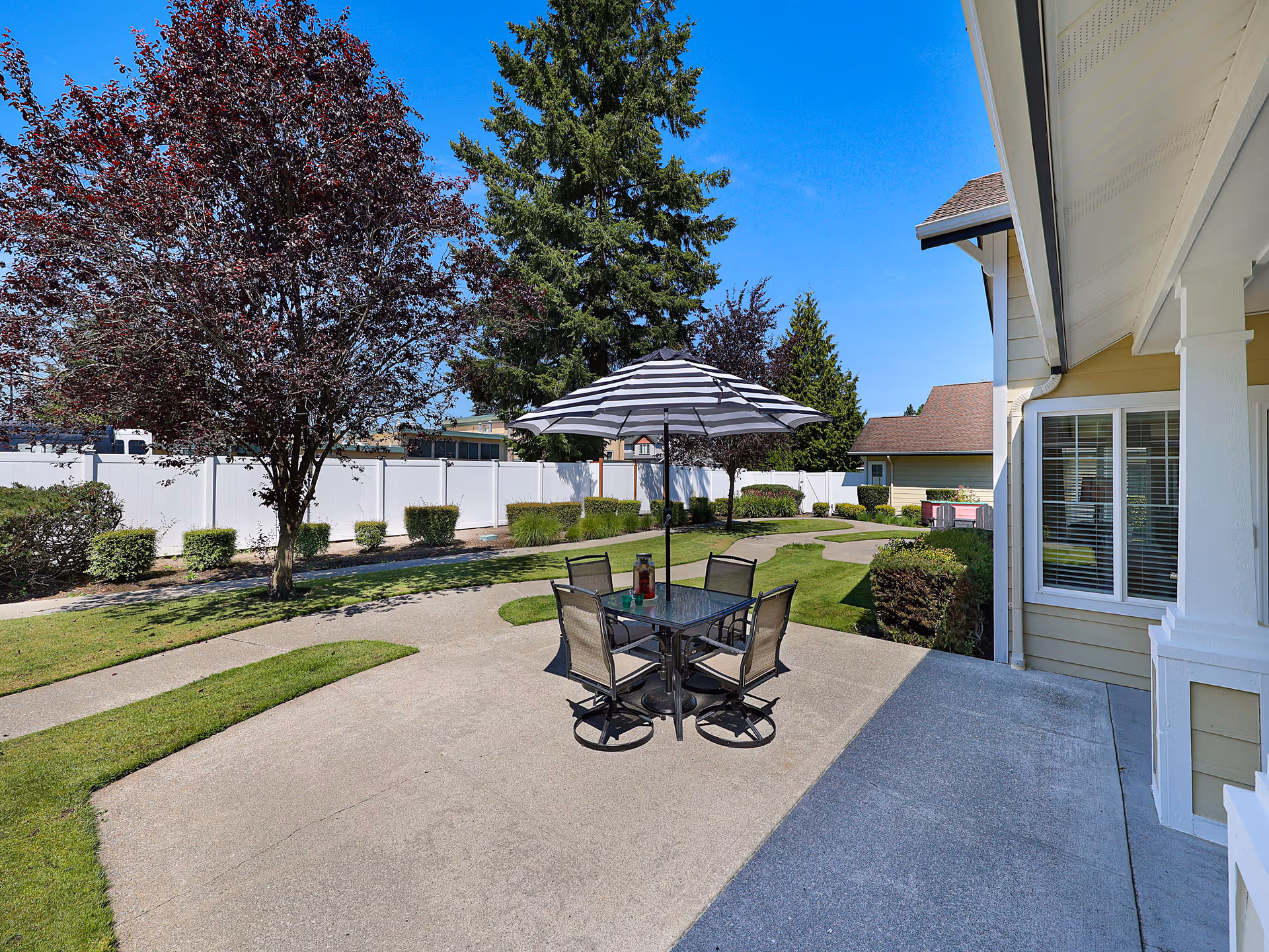Outdoor patio area at White River Memory Care featuring a glass table with four chairs and a striped umbrella. The patio is surrounded by green grass, bushes, and trees with a clear blue sky overhead. A building with windows and a covered porch is visible on the right side.