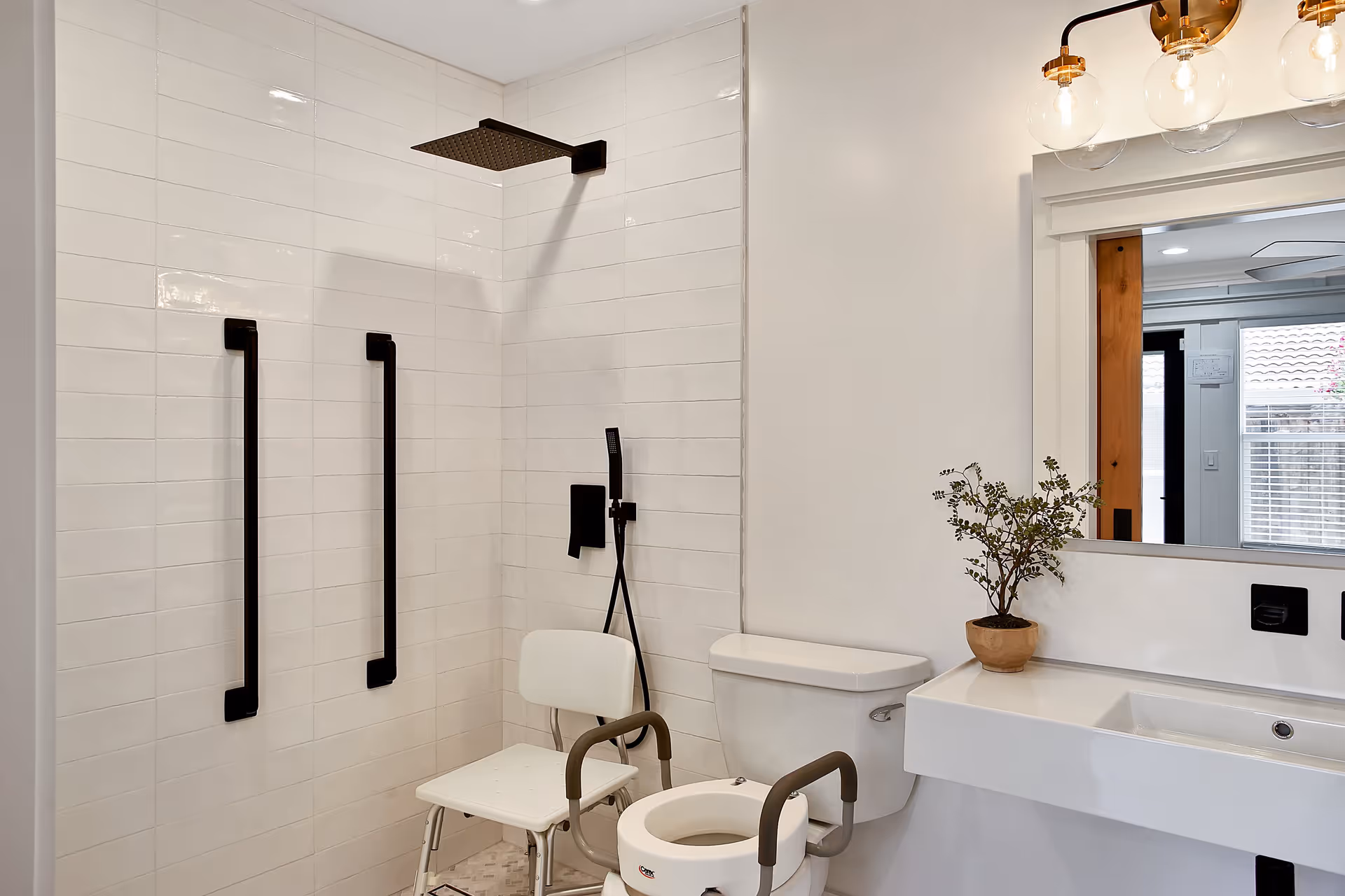 A modern senior-friendly bathroom featuring a walk-in shower with two black grab bars, a white shower chair, and a black showerhead. There is a toilet with padded arm supports, a white sink with a potted plant on the counter, and a large mirror reflecting a window with blinds.