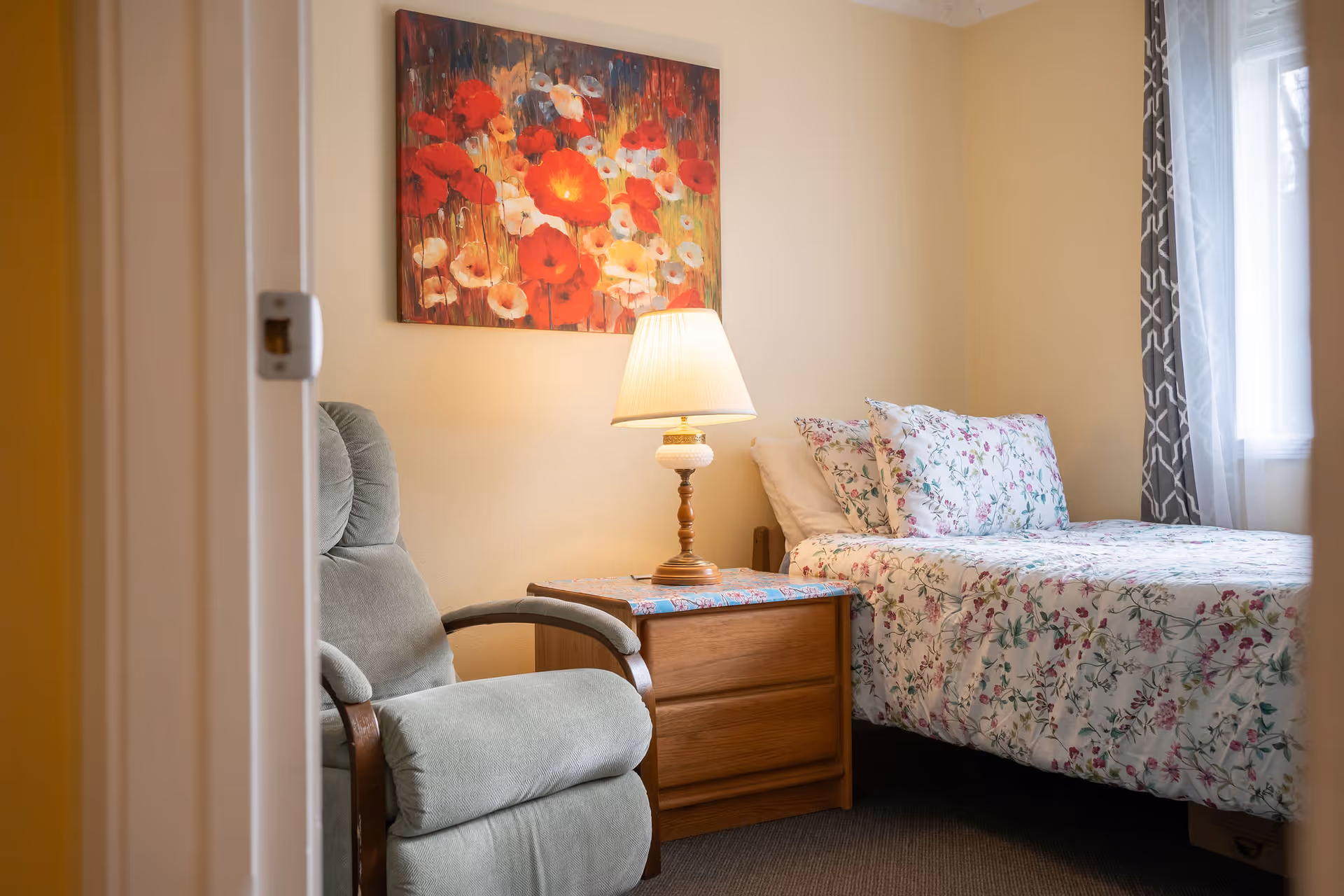 A cozy bedroom with a floral-patterned bedspread and matching pillows on a bed next to a wooden nightstand with a lamp. A comfortable green armchair is positioned beside the nightstand. A colorful painting of red and white flowers hangs on the beige wall above the armchair. Light filters in through a window with patterned curtains.