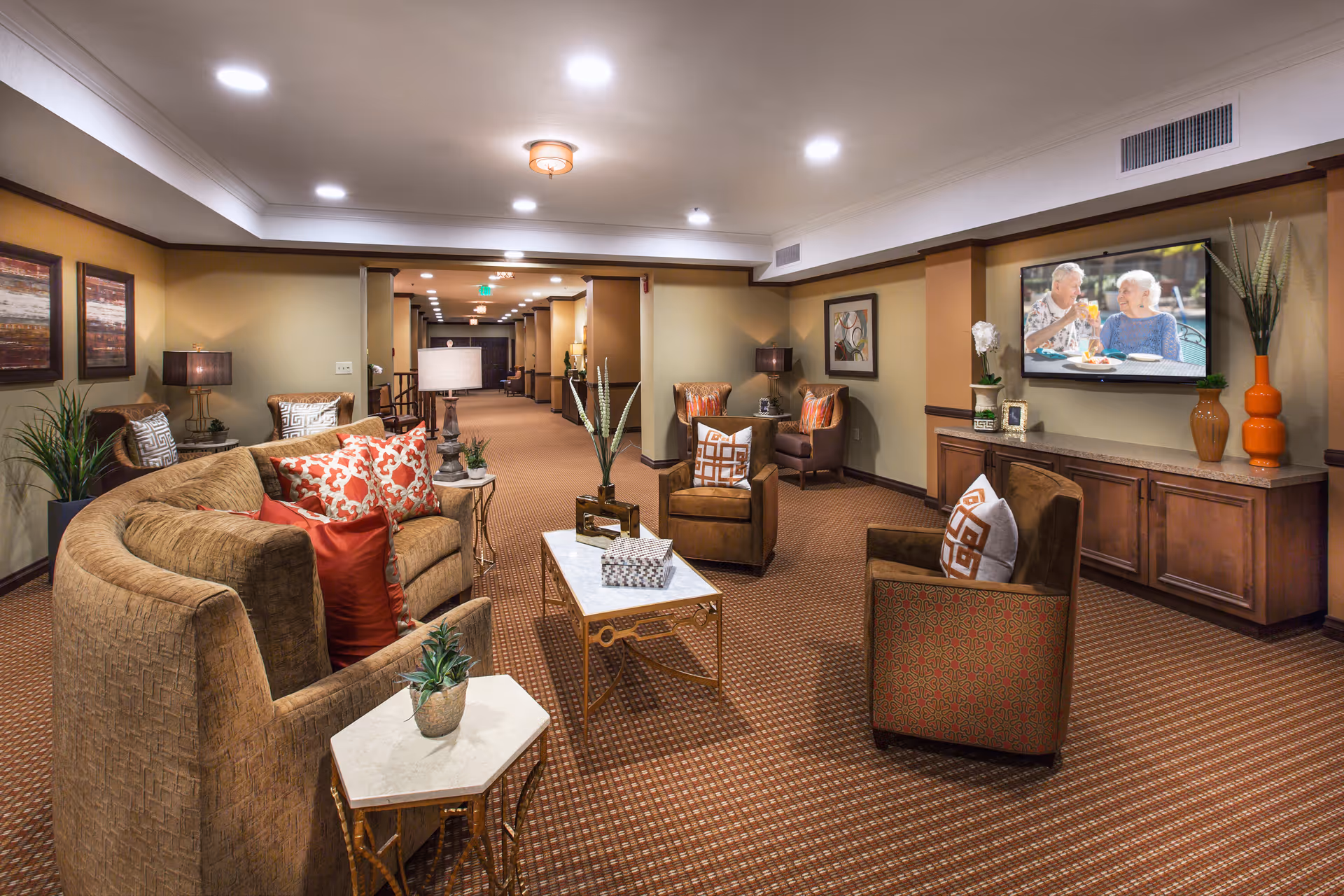 Comfortable communal lounge with sofas, armchairs, coffee tables and a wall-mounted TV in a senior living facility.