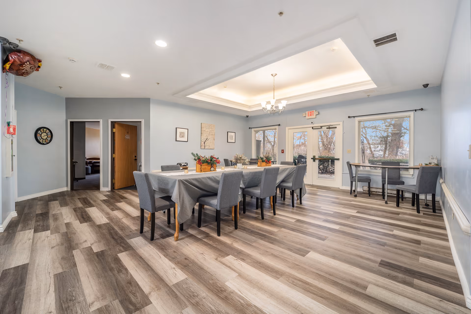 Bright communal dining room with a long table covered in a gray tablecloth, upholstered chairs, chandelier, and double doors to the outside.