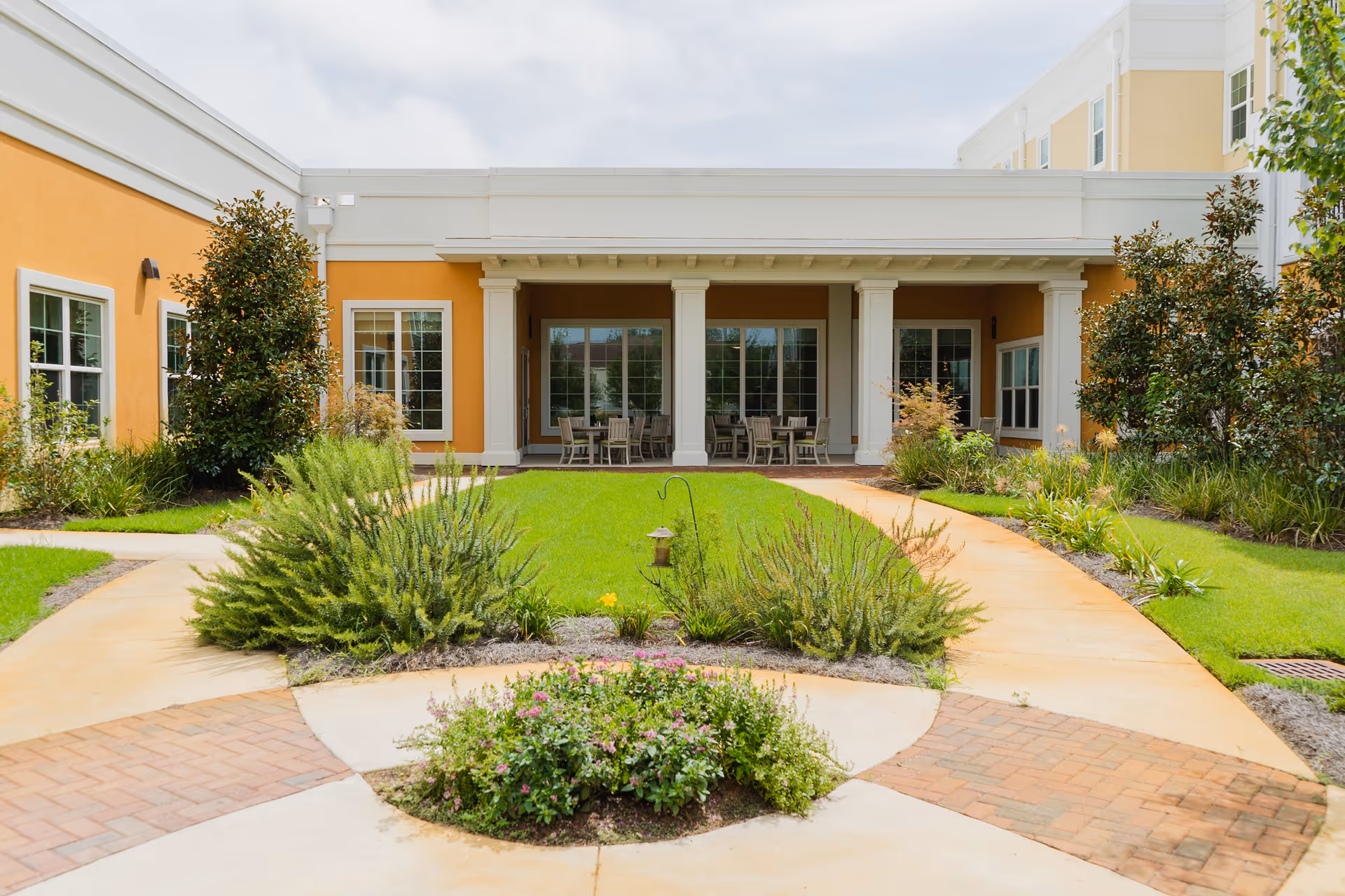 Landscaped courtyard with paved paths leading to a covered patio with columns and outdoor seating at a senior living building.