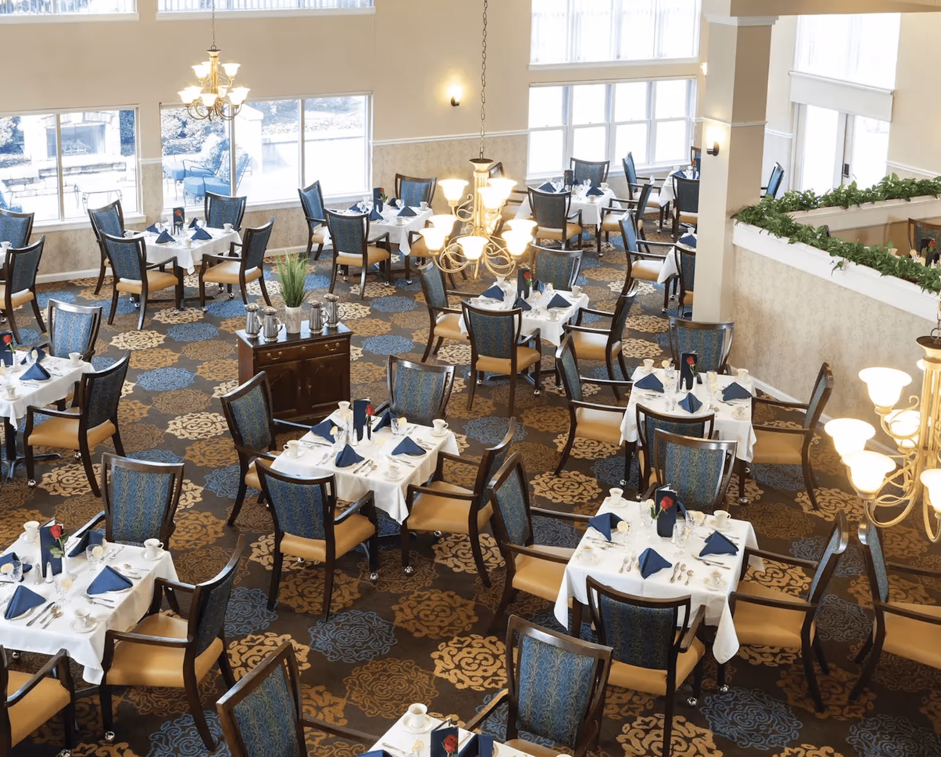 A spacious dining room with multiple tables covered in white tablecloths, each set with blue napkins, cups, and silverware. The room features large windows allowing natural light to fill the space, patterned carpet in shades of blue and brown, and elegant chandeliers hanging from the ceiling. Chairs with blue patterned backs and tan seats surround each table.