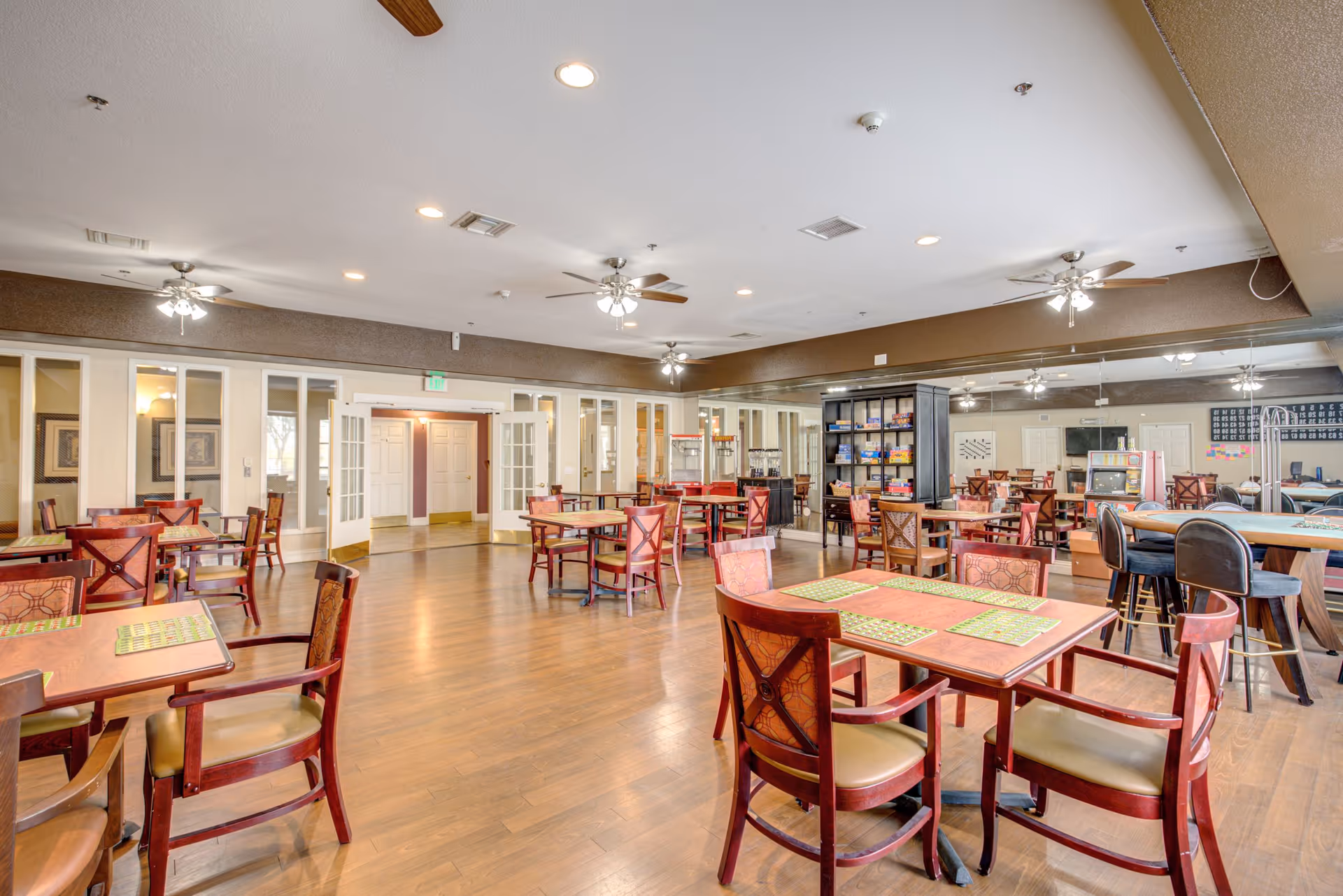 A spacious community room with multiple wooden tables and chairs arranged neatly on a wooden floor. The room features ceiling fans with lights, a large mirror on one wall, and a black shelving unit stocked with board games and snacks. There is also a popcorn machine and a card table with chairs, creating a welcoming and social atmosphere.