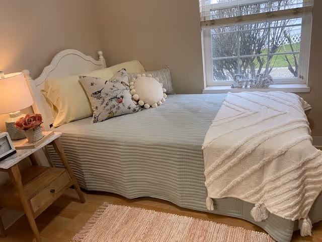 A cozy bedroom with a neatly made bed featuring a light-colored quilt, decorative pillows including a round pom-pom pillow, and a textured throw blanket. Next to the bed is a wooden nightstand with a lamp, a small flower arrangement, and a framed photo. A window with white blinds allows natural light to brighten the room, showing a view of trees and grass outside.