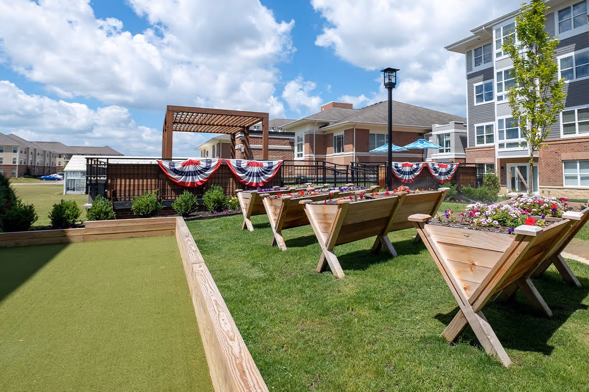 Outdoor garden area at Verena at Hilliard featuring raised wooden flower beds with colorful flowers, a bocce ball court with green turf, a pergola, and a multi-story residential building in the background under a partly cloudy sky.