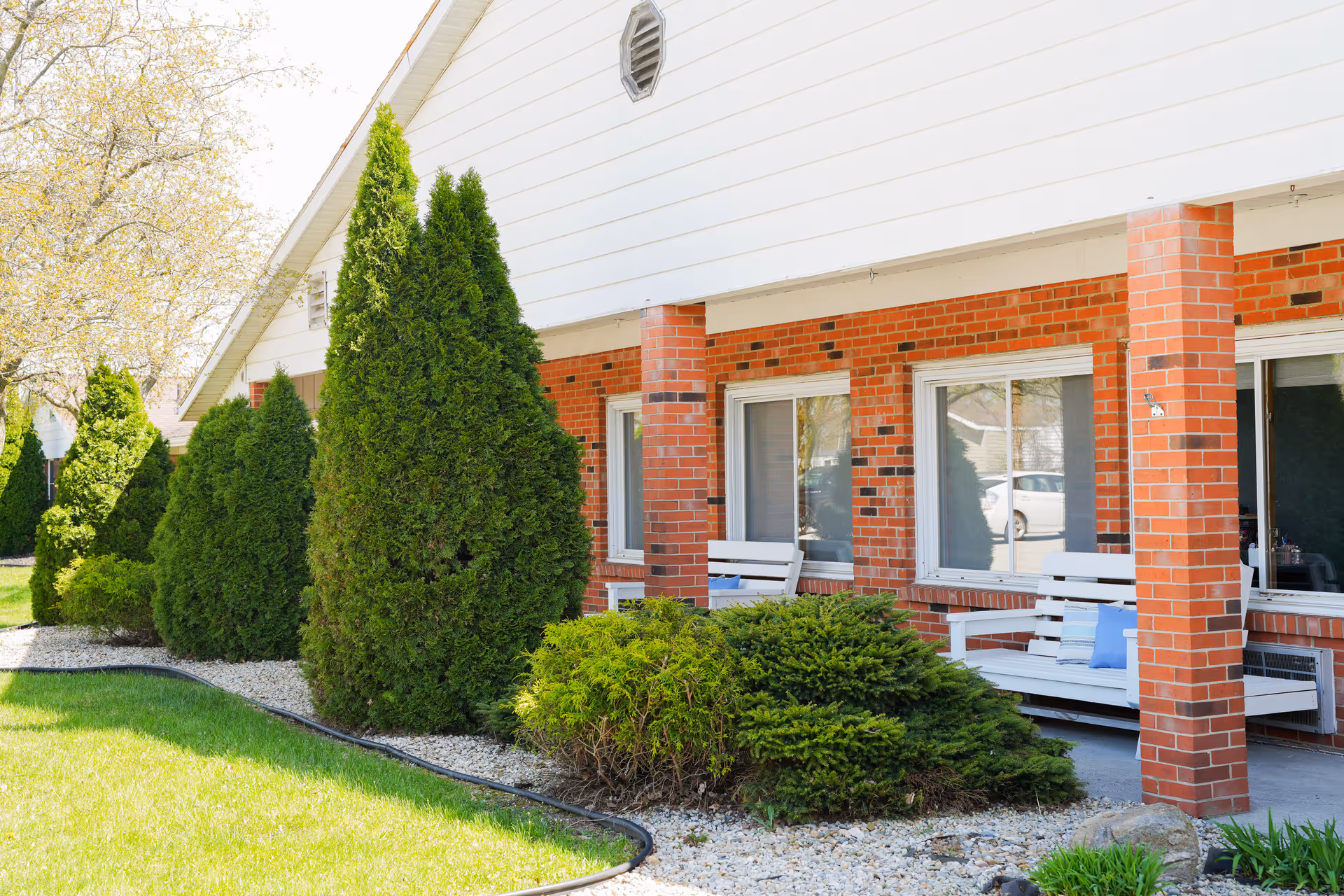 Exterior view of a senior living facility with a brick facade and white siding. The building has a covered porch area with white benches and blue cushions. There are several tall green shrubs and bushes planted along the front, with a well-maintained lawn and trees in the background.