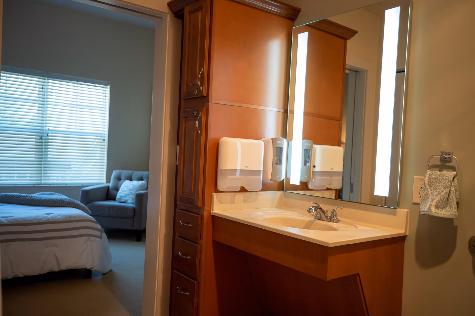 Vanity sink with illuminated mirror, soap dispensers, and towel beside an open doorway showing a bedroom with a bed and armchair.