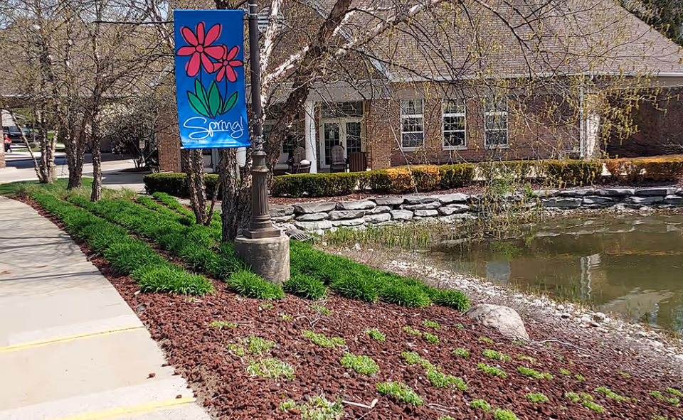 Exterior view of a brick building with a sidewalk, landscaped beds, a lamp post displaying a 'Spring!' banner, and a small pond.